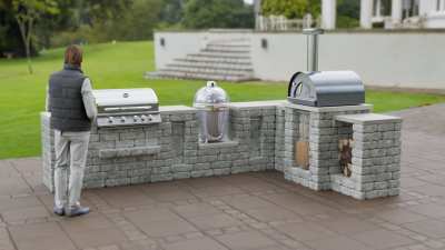 Man on patio using outdoor kitchen with pizza oven, gas grill and. egg smoker.